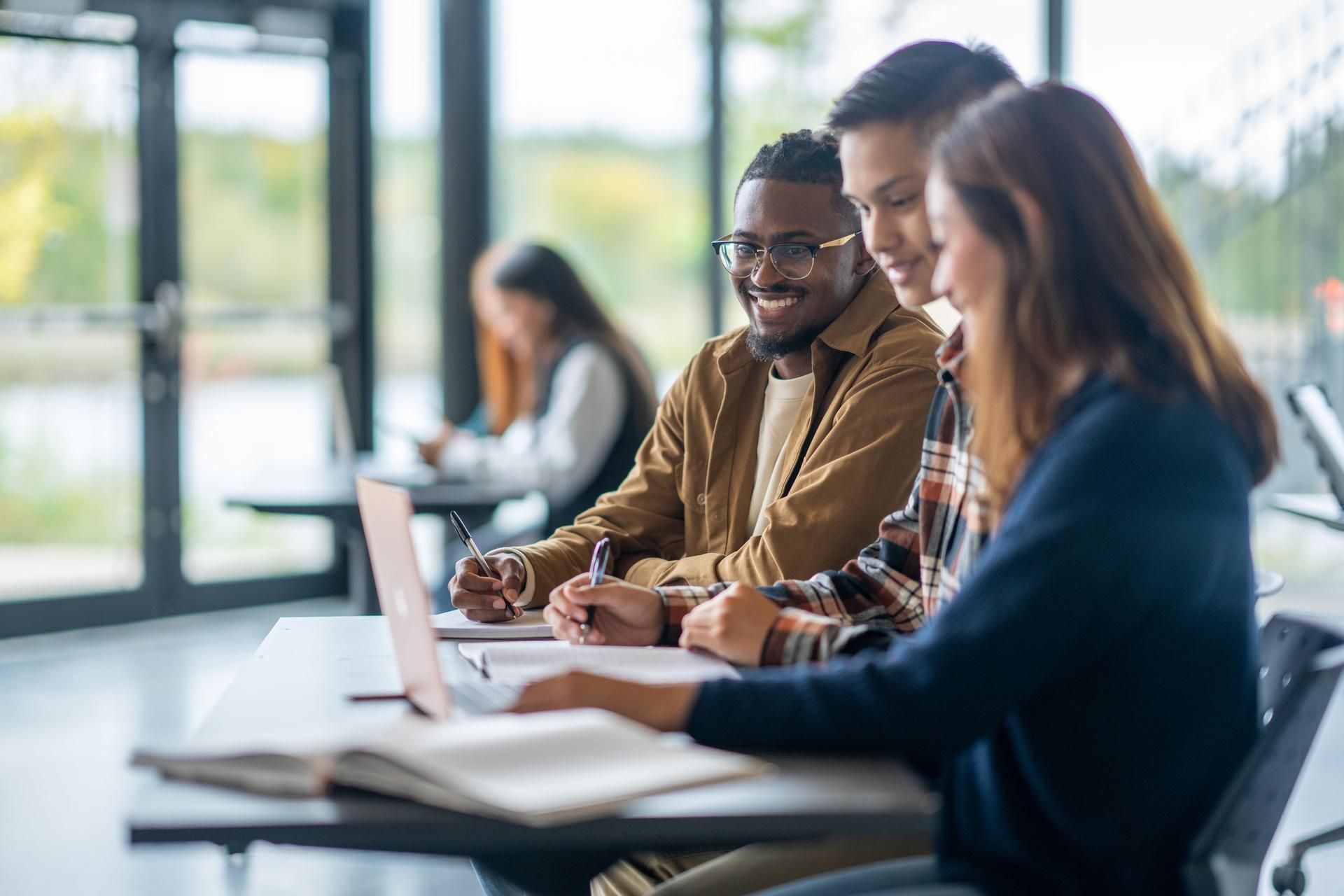 A group of three students sits around a table, working intently on a laptop, sharing ideas and knowledge.