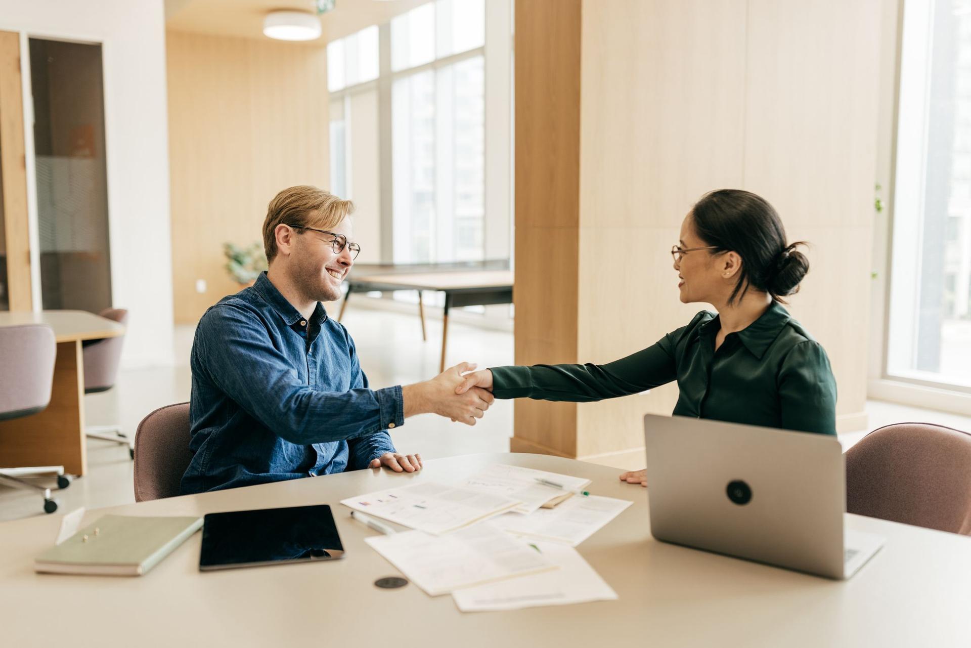 Two people engaged in a handshake at a table in an office, representing a mutual agreement.