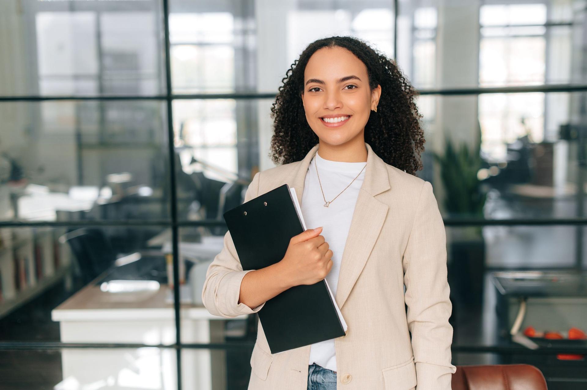 A confident young teacher smiles while holding a folder in a bright school setting.