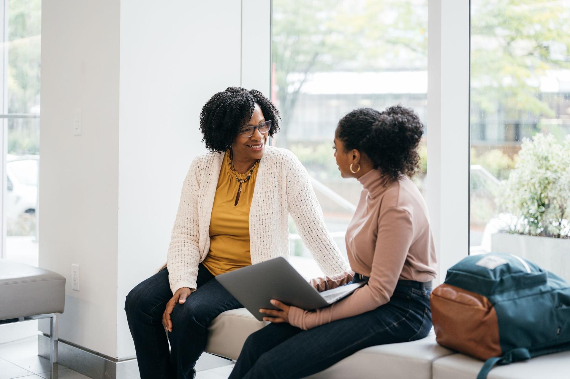 Two women, a professor and a student, engaged in conversation while sitting on a bench at a college campus.