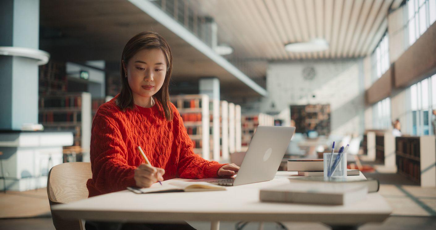 A focused female student sits at a table in a library, writing on her laptop amidst a quiet study environment.