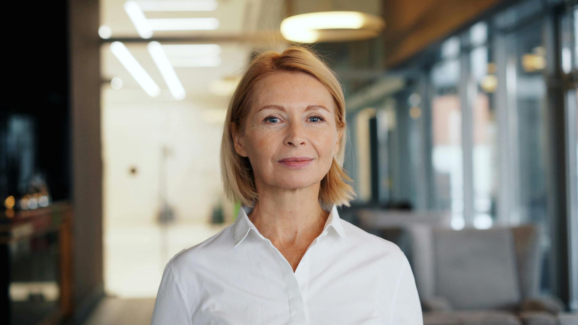 A woman with short blonde hair and a white shirt stands smiling in a well-lit, modern office setting.