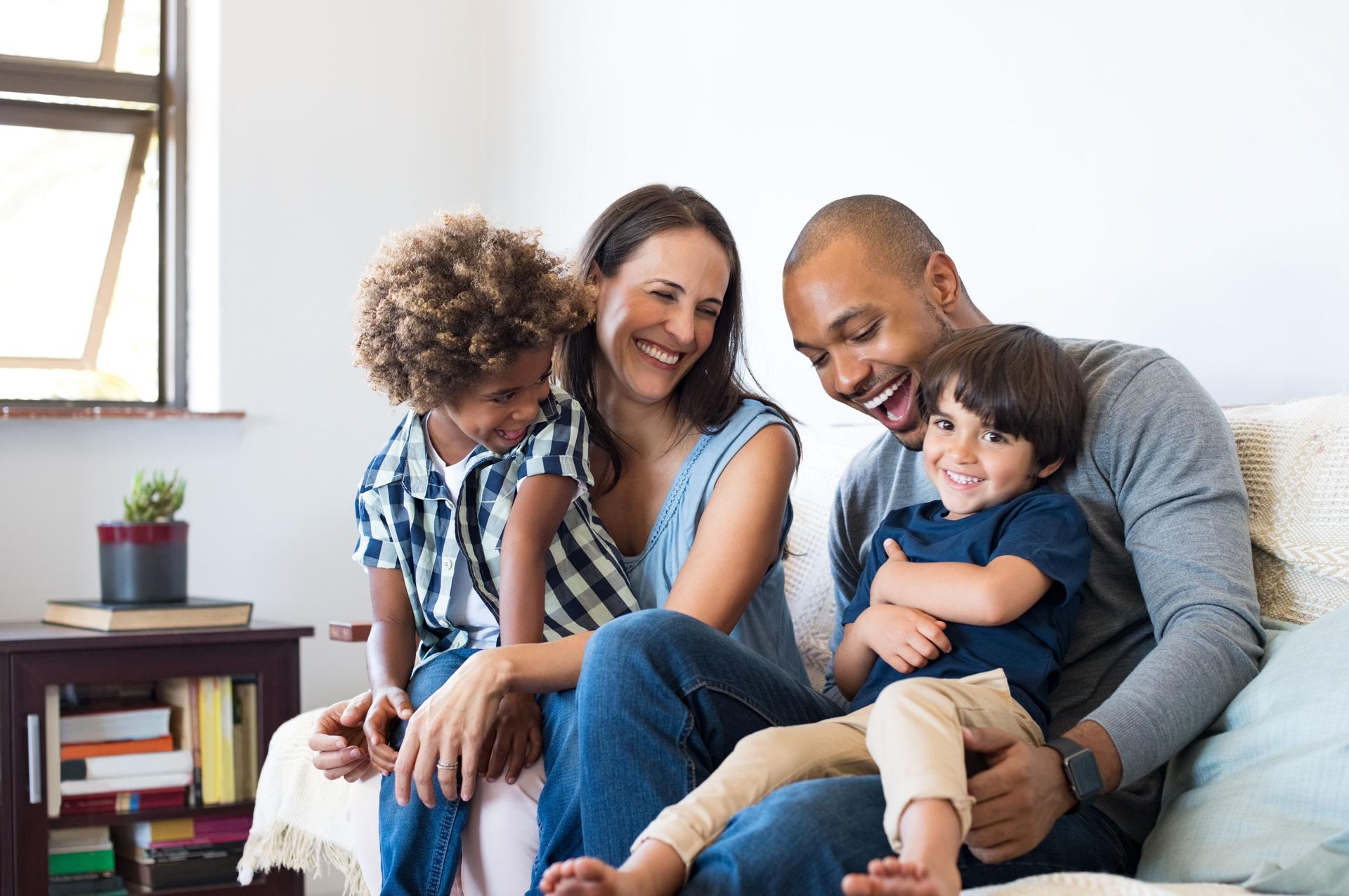 Happy family sitting on a couch together, including a smiling father, mother, and two young children, one with curly hair and the other with straight hair, enjoying a playful moment in a bright, cozy living room.