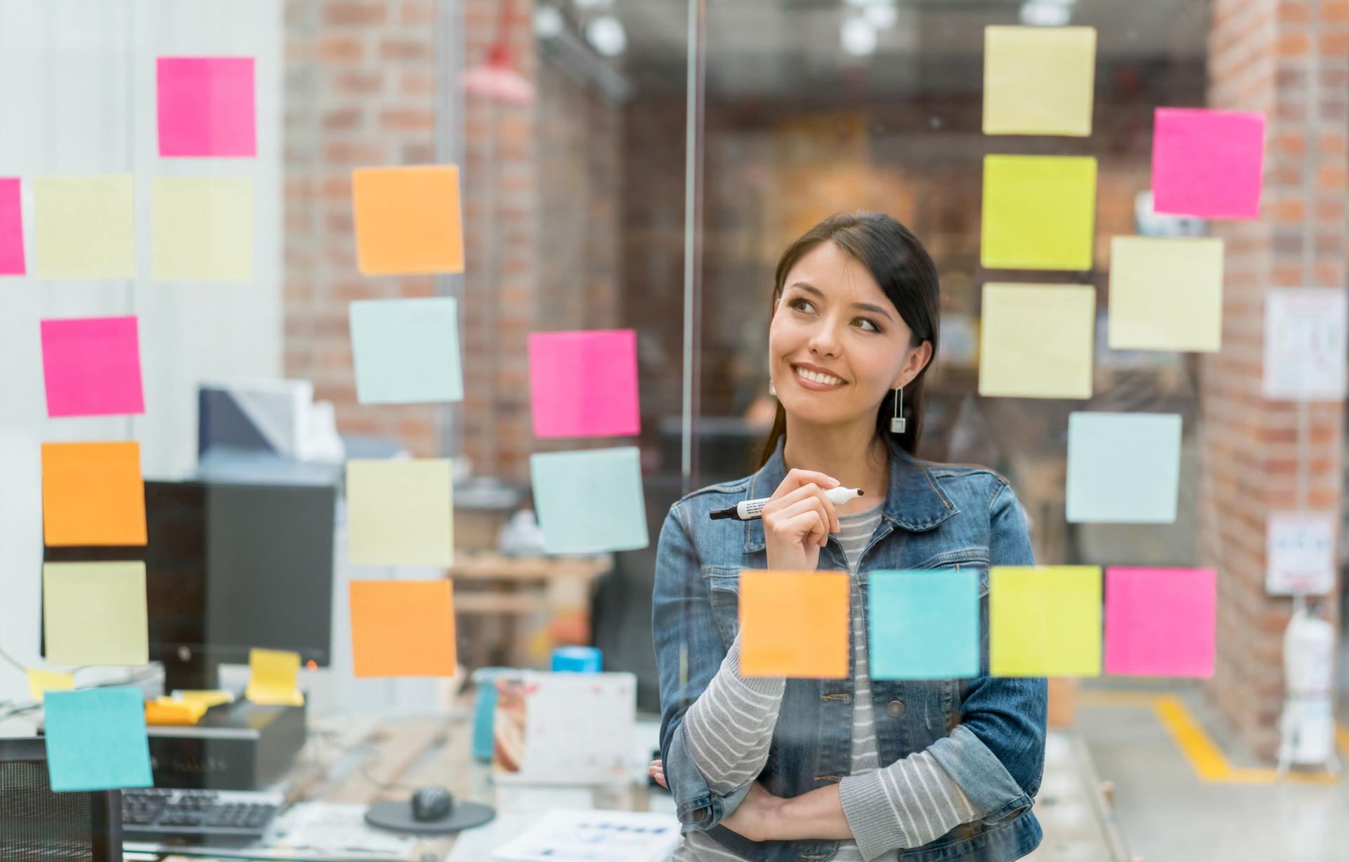 A student stands thoughtfully in front of a glass wall covered in colorful sticky notes, contemplating her ideas.