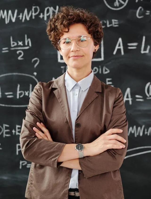 A confident person with short curly hair and glasses stands with arms crossed in front of a blackboard filled with mathematical equations. They are wearing a brown blazer over a light blue shirt.