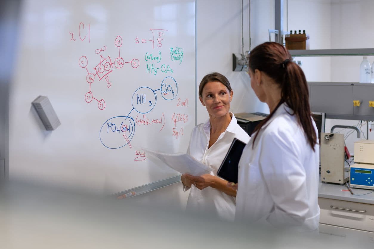 Two women wearing lab coats are positioned next to a whiteboard, engaged in a discussion about scientific concepts.