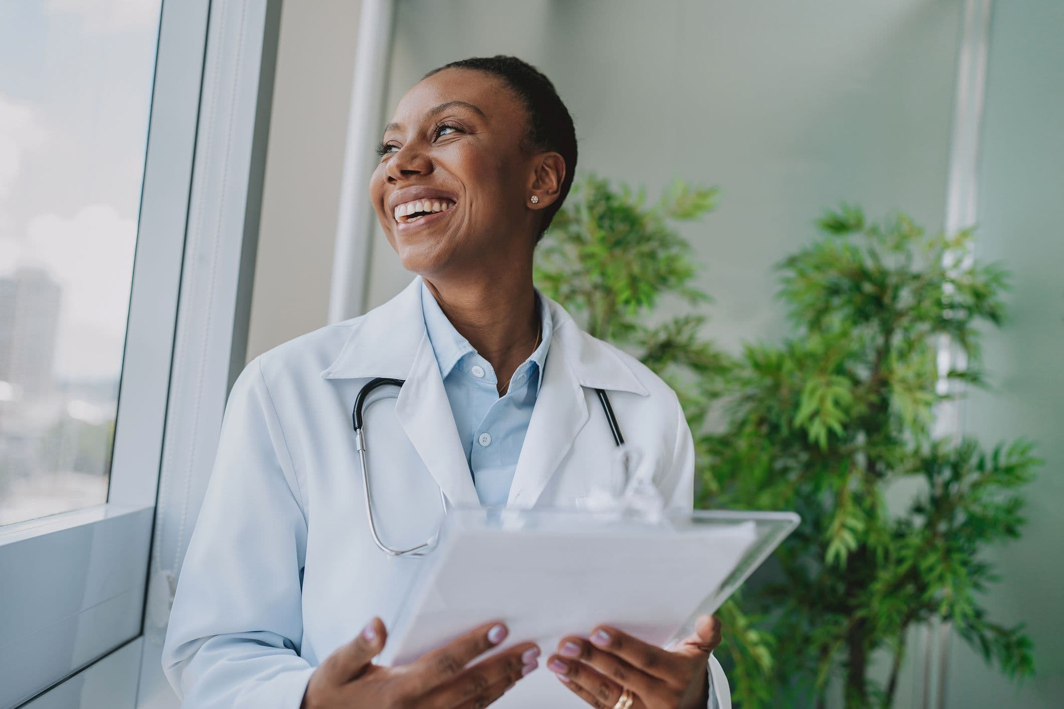 A Black female doctor in a white coat smiles while holding a clipboard, symbolizing her role in health care.