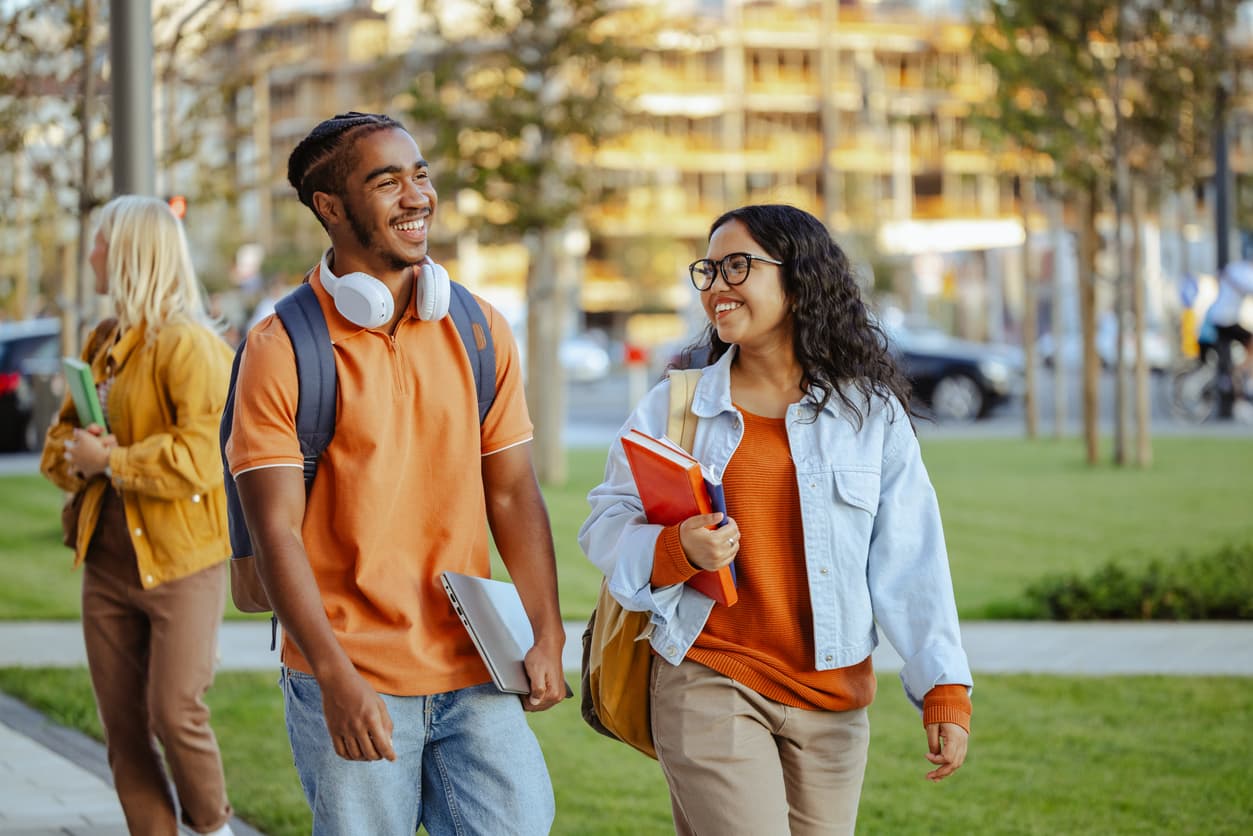 Two young students walk on a sidewalk, each carrying books under their arms, engaged in conversation.