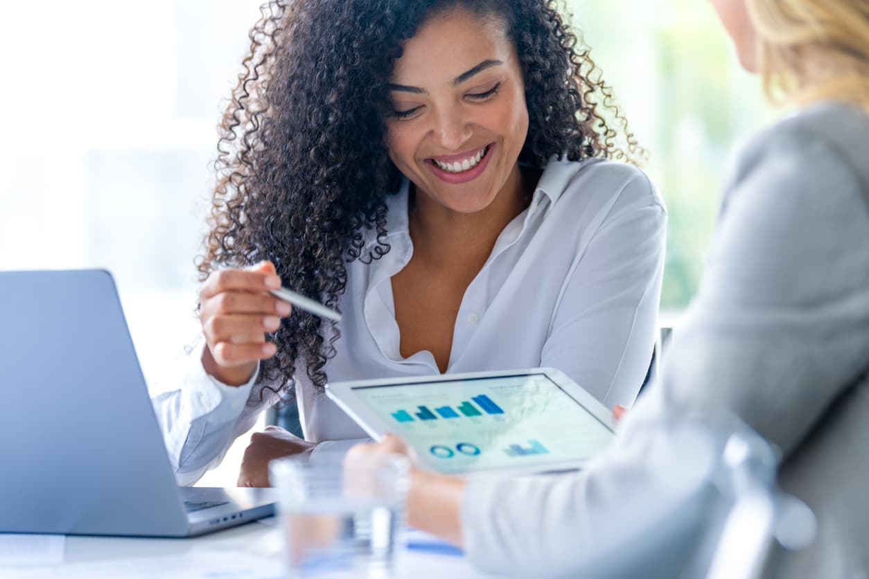 Two women sit at a table, focused on a laptop, engaged in discussion or collaboration.
