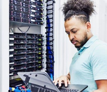 A person with curly hair tied up is standing in a server room, working on a laptop. They are focused on the screen, with several server racks and network cables visible in the background.