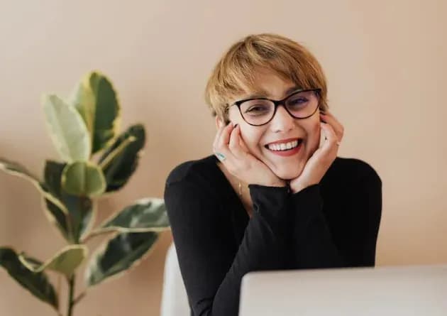 A smiling academic advisor with short blonde hair and glasses leans on her hands in front of a laptop. The background features a plant, creating a warm and welcoming environment.