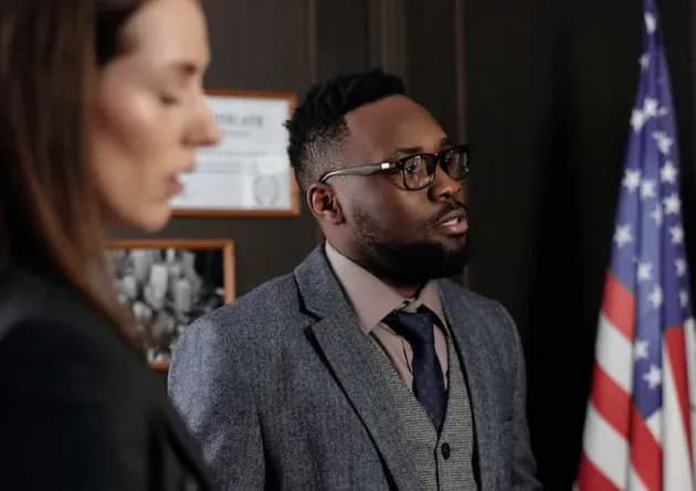 A man and woman in a formal setting, possibly a courtroom or government office. The man, wearing glasses, a suit jacket, and a tie, speaks seriously. An American flag and framed documents are visible in the background, indicating a professional environment.