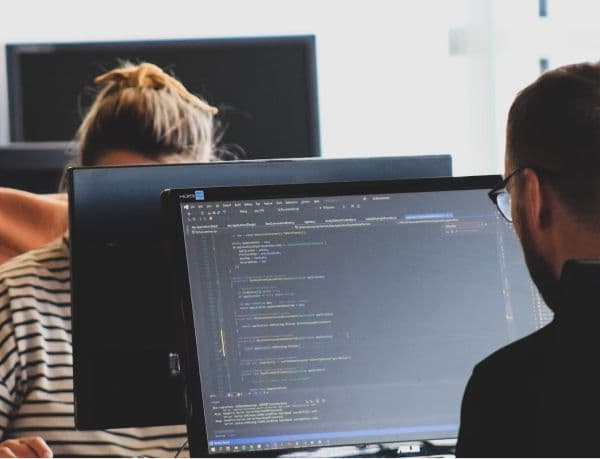 Two people work at their desks, focused on computer screens displaying code. One person wears a striped shirt, the other glasses. The setting is a collaborative office environment, suggesting software development or data analysis.