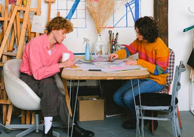 Two people are sitting at a round table in an art studio, engaged in a creative project. The person on the left has curly hair, wears a bright pink shirt, and holds a coffee cup. The person on the right has dark curly hair, wears a colorful striped sweater, and is focused on the project. The table is covered with papers and art supplies. The background features easels, drawings, and various art materials, indicating a busy and vibrant creative environment.