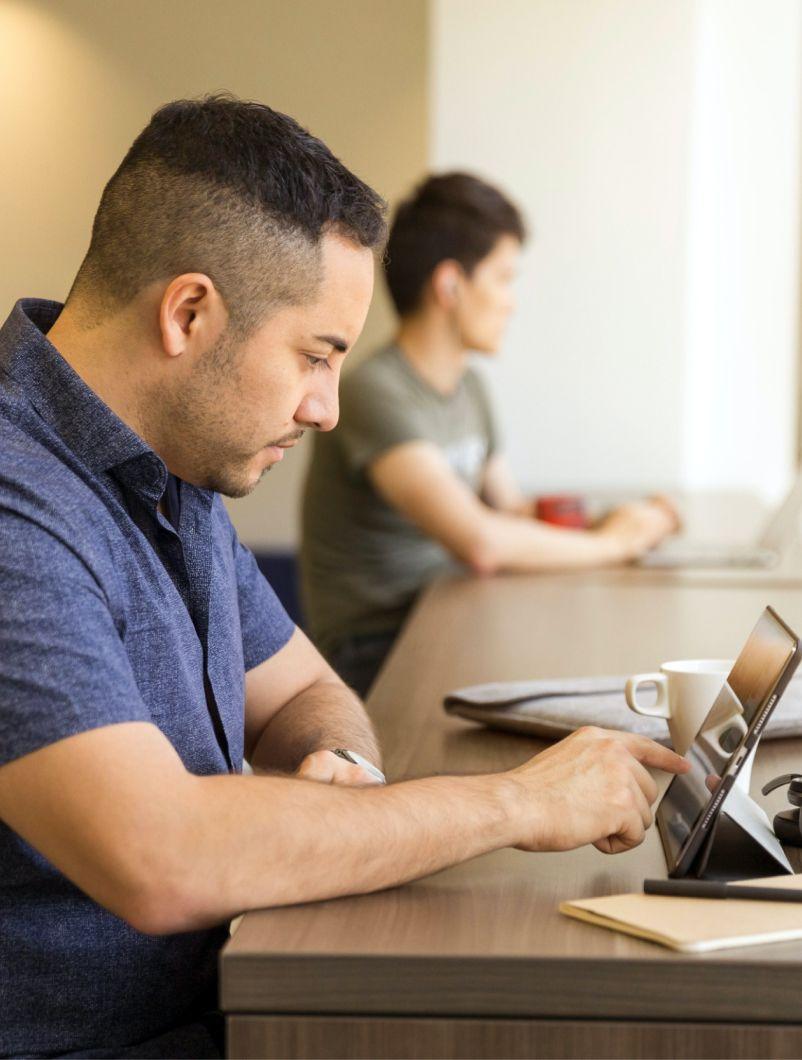 A man with short dark hair is seated at a long table, focused on working with a digital tablet. He is wearing a blue shirt and is engaged in his task. In the background, another person is also working on a laptop. The setting appears to be a quiet, modern workspace, with a cup of coffee and a notepad on the table, creating a studious and productive environment.