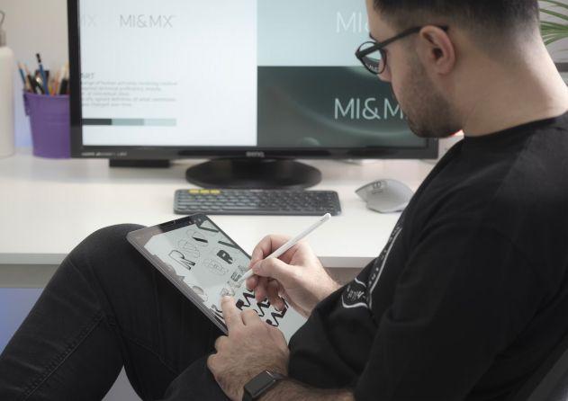 A man with short dark hair and glasses is working on a digital drawing tablet at a modern white desk. He is focused on sketching designs, with a large monitor displaying "MI & MX" in front of him. The organized desk includes a keyboard, mouse, and a purple container with art supplies.