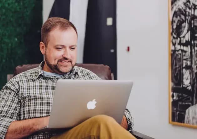 A smiling assessment and testing developer works on a laptop in a relaxed setting. The image captures the professional in a moment of focused engagement with his work.