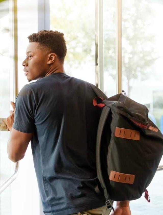 A person with short hair is standing near a glass door, looking to the side. They are wearing a dark t-shirt and have a large black backpack with brown accents slung over one shoulder. The background is bright, with light streaming through the glass and a blurred view of the outdoors.