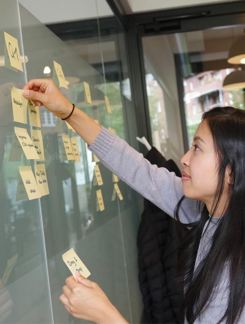 A woman with long dark hair is placing sticky notes on a glass wall. She is wearing a grey sweater and appears to be organizing tasks or ideas. The background shows a modern office space with large windows and a view of buildings outside. The sticky notes have various writings on them, indicating a planning or brainstorming session. The atmosphere suggests a focused and collaborative work environment.