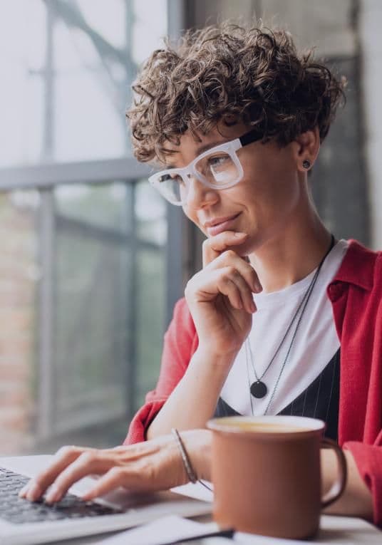 A person with short curly hair and wearing glasses is sitting at a desk, thoughtfully looking at a laptop screen. They are wearing a red cardigan over a white shirt and have a necklace with a round pendant. A cup of coffee is placed on the desk in front of them. The background shows a large window with a blurred outdoor view.