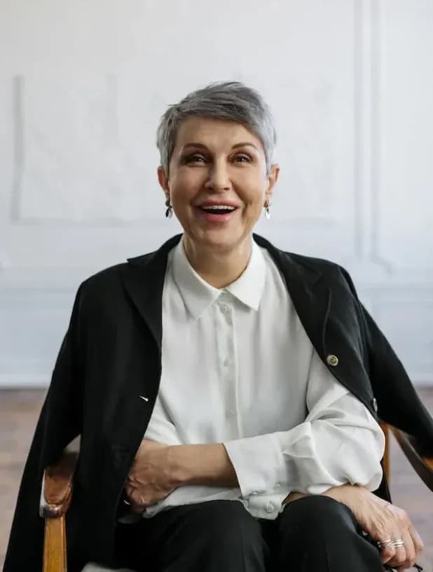 A smiling advisor with short gray hair sits confidently in a chair. She is dressed in a white blouse and a black jacket, exuding professionalism and approachability.