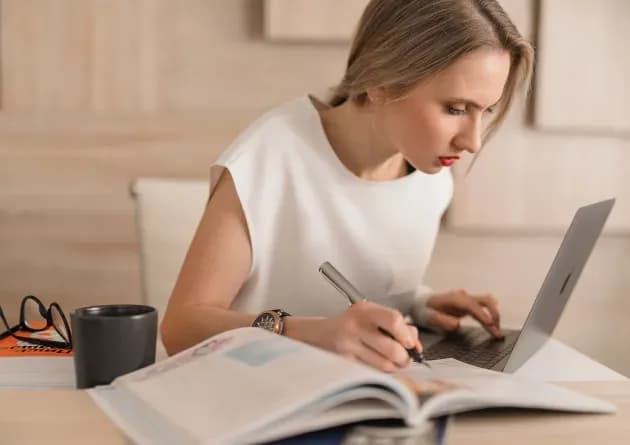 A focused curriculum developer works on a laptop while taking notes from an open book. The scene conveys dedication and attention to detail in creating educational materials.