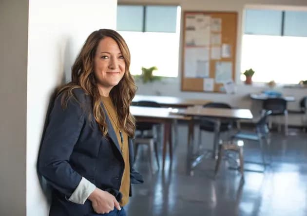 A smiling education consultant leans against a wall in a classroom setting. She is dressed in professional attire, exuding confidence and expertise in educational consulting.