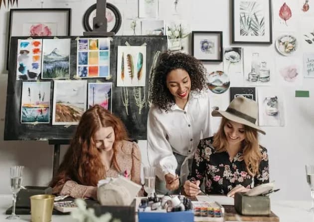 An elective art teacher helps two students with their paintings in a creative studio. The background is filled with various art pieces, creating an inspiring and artistic environment.