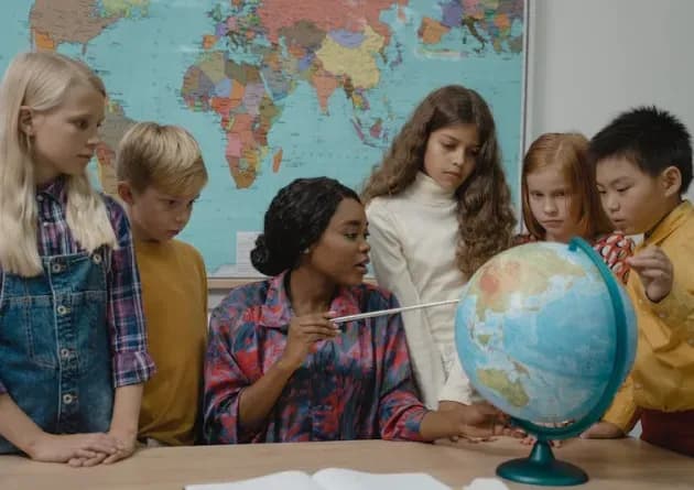 An elementary school teacher points at a globe while teaching a group of attentive children. They are gathered around a desk with a world map in the background, creating an engaging learning environment.