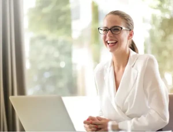 Confident executive woman in a white blazer, smiling while working on a laptop in a bright, modern office environment.