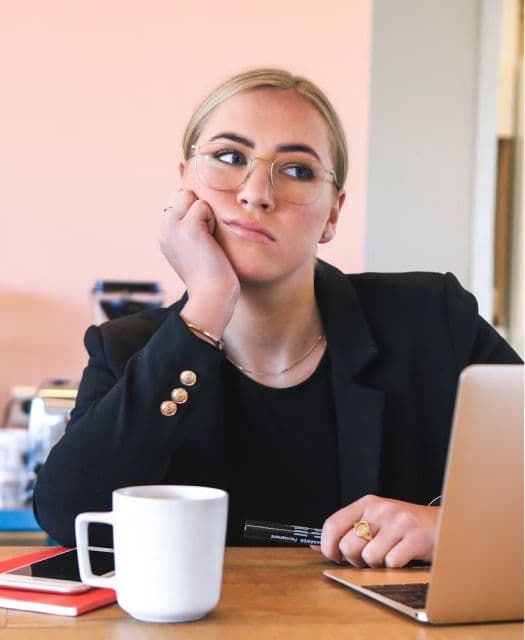 A woman in business attire sits at a desk, looking contemplative. She rests her chin on her hand, with a laptop and coffee mug in front of her. She appears to be deep in thought, perhaps pondering her career path or next steps.