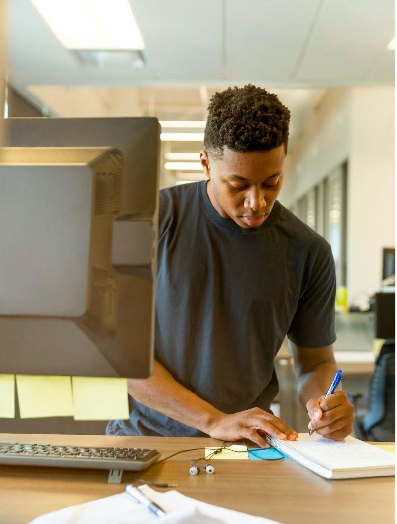 A focused student is working at a desk in a modern study environment. He is writing in a notebook while standing, with a computer monitor and keyboard in front of him. The setting appears to be a well-lit office or study space, indicating a productive and organized workspace.