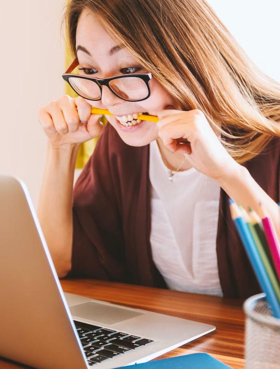 A woman biting a pencil in front of a laptop.