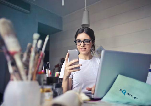 A young woman with dark hair and glasses is sitting at a cluttered desk, holding a smartphone and smiling. She is wearing a white t-shirt and working on a laptop. The desk is filled with various art supplies, including brushes, pens, and containers, suggesting she is in a creative workspace. A soft light illuminates the room, creating a productive and engaging atmosphere.