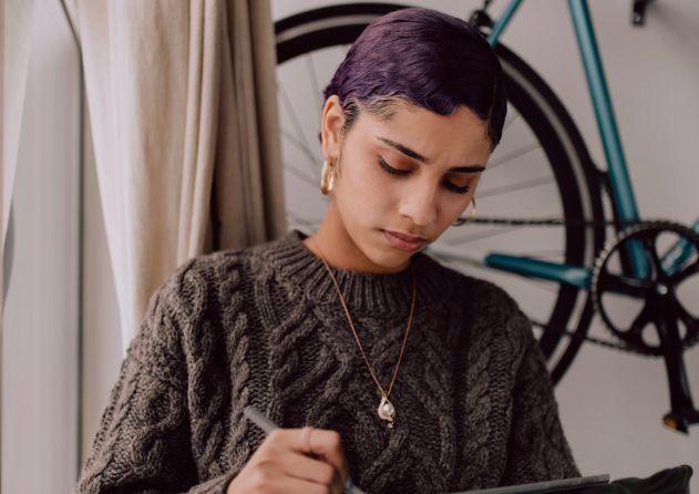 A young woman with short, purple hair is focused on her work, writing or drawing on a tablet. She is wearing a textured, dark sweater and a necklace with a pendant. The background features a bicycle mounted on the wall and a window with curtains, suggesting a creative and cozy workspace.