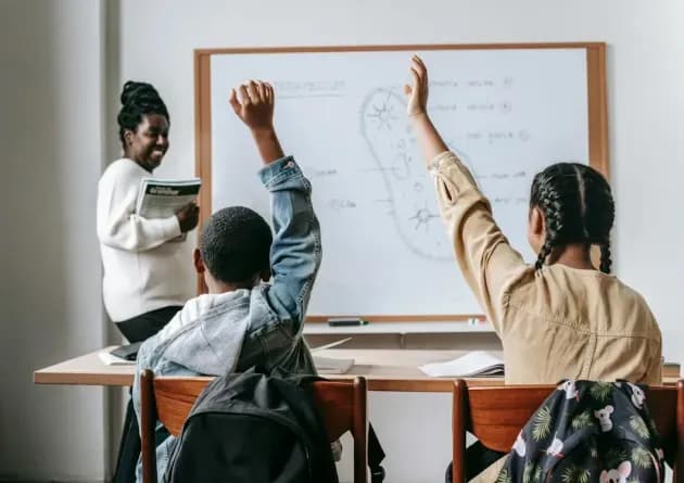 A high school teacher smiles as two students raise their hands to answer a question during a biology lesson. The whiteboard behind them shows a diagram and notes, creating an interactive classroom environment.