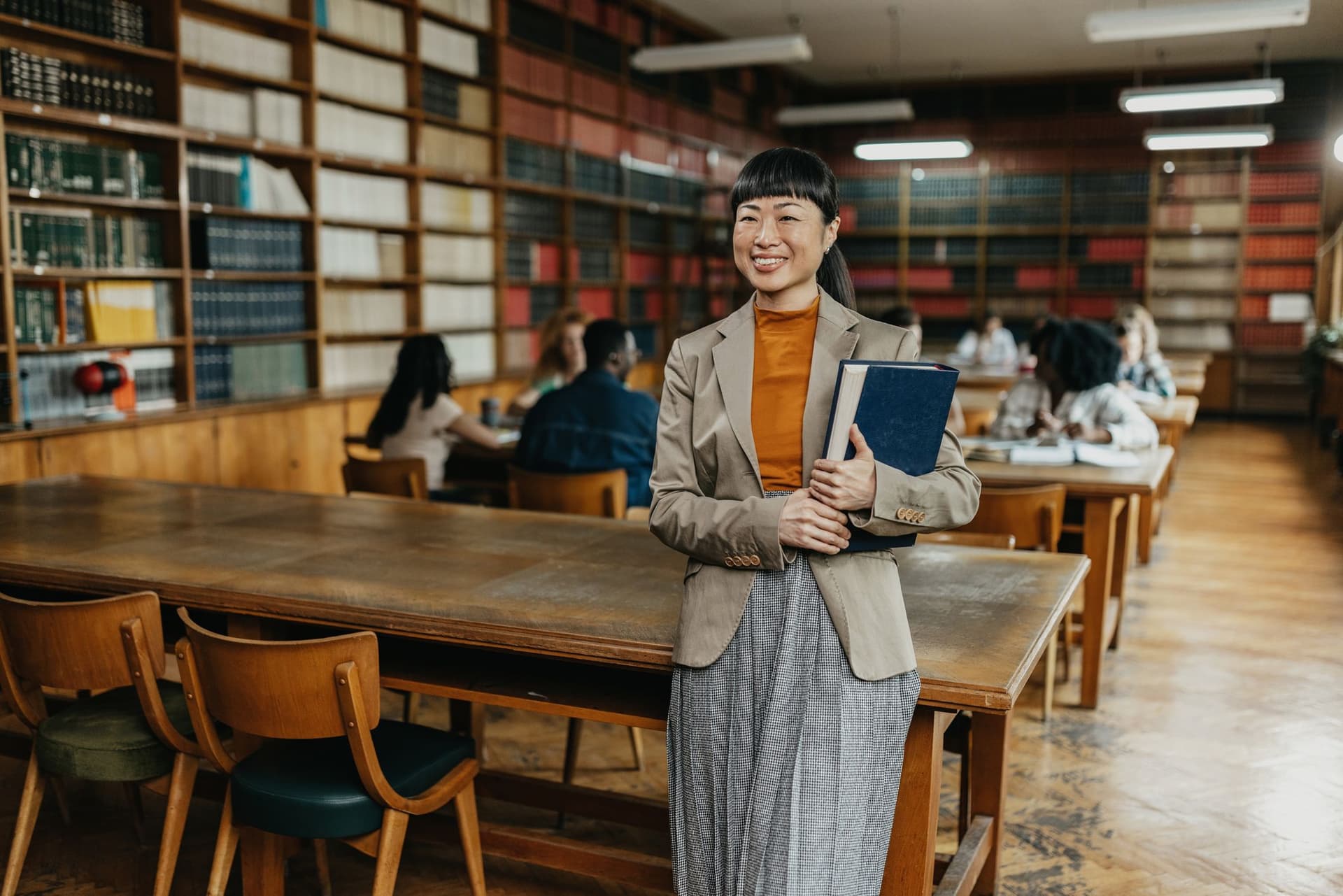 A teacher in a library holds a book, surrounded by shelves filled with various books.