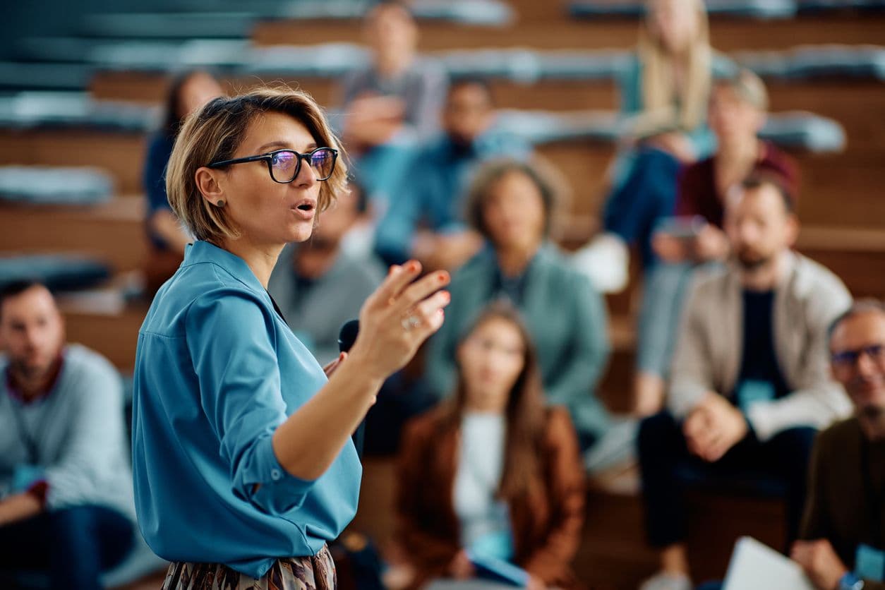 A woman with glasses stands before an audience, presenting educational material to her fellow students.