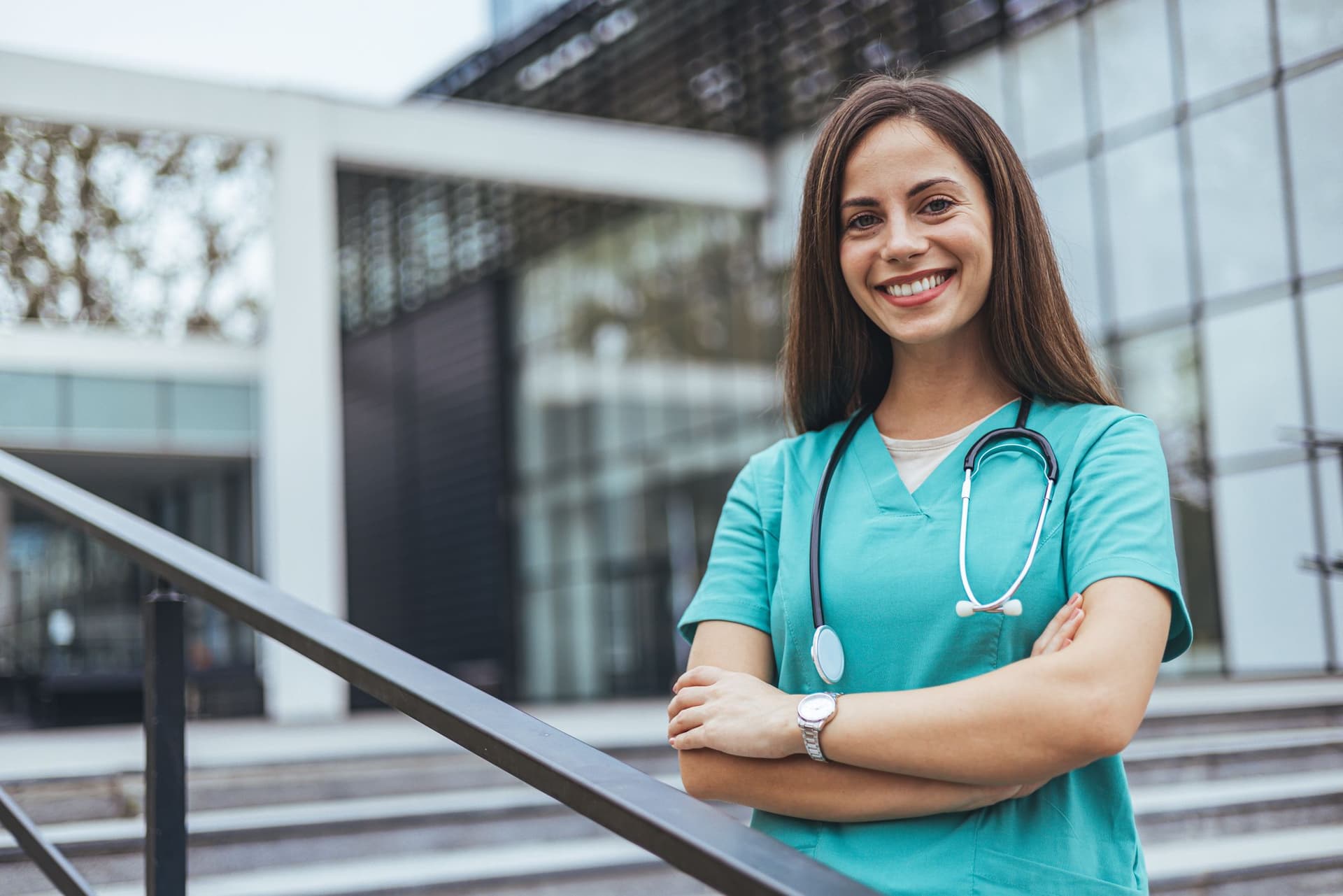 A female nurse stands on a stairway, dressed in scrubs, with a stethoscope around her neck, appearing professional and caring.
