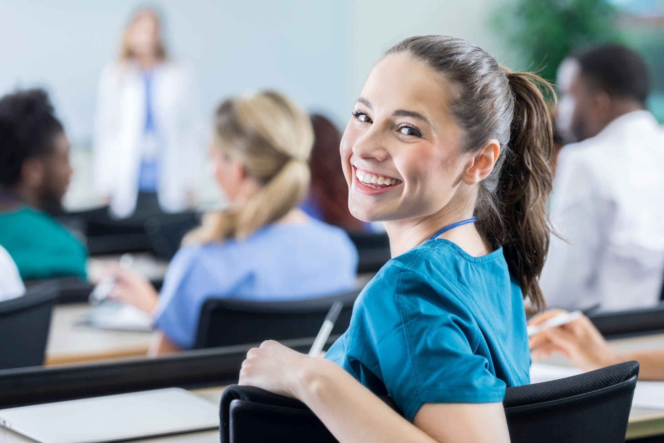 A smiling woman in scrubs sits in a classroom, facing the camera with a welcoming expression.