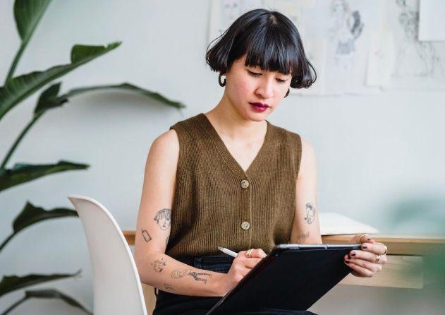 A woman with short black hair and tattoos is sitting at a desk, focused on working with a digital tablet and stylus. She is wearing a brown sleeveless top and black pants. The background includes a large plant and sketches pinned on the wall, suggesting a creative and artistic workspace. The atmosphere is calm and professional, emphasizing her engagement in the design work.