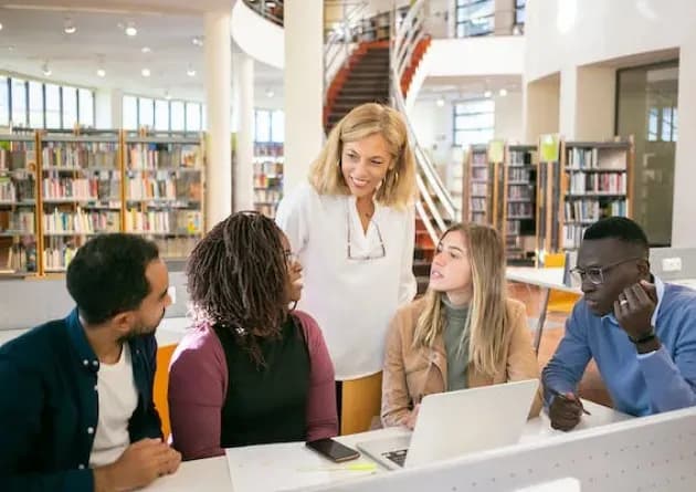 A librarian assists a diverse group of students gathered around a laptop in a library. The setting is bright and spacious, filled with bookshelves, indicating a collaborative and supportive learning environment.