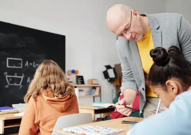 A middle school teacher in a classroom assisting a student with their work. Other students are focused on their tasks, with a blackboard displaying math problems in the background, creating an active learning environment.