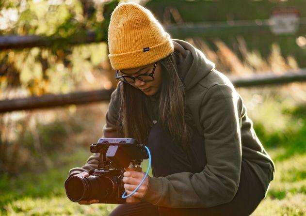 A woman wearing a yellow beanie, glasses, and a green hoodie is crouching outdoors, focused on adjusting a professional camera. The background is a natural setting with sunlight filtering through, highlighting the grassy area and wooden fence behind her. She appears to be engaged in capturing a shot, indicating a moment of creative work in a serene environment.