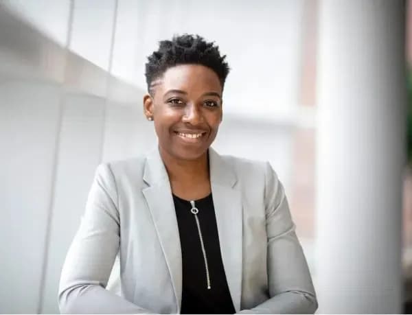 A professional woman wearing a light gray blazer and a black top, smiling confidently while standing in a bright, modern setting.