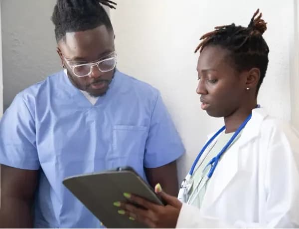 Two healthcare professionals, one in blue scrubs and the other in a white lab coat with a stethoscope, reviewing information on a tablet.