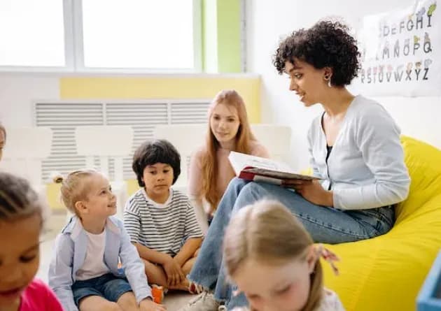 A preschool teacher reads a book to a group of young children seated on the floor in a classroom. Another adult is also present, engaging with the children, creating a warm and interactive learning environment.