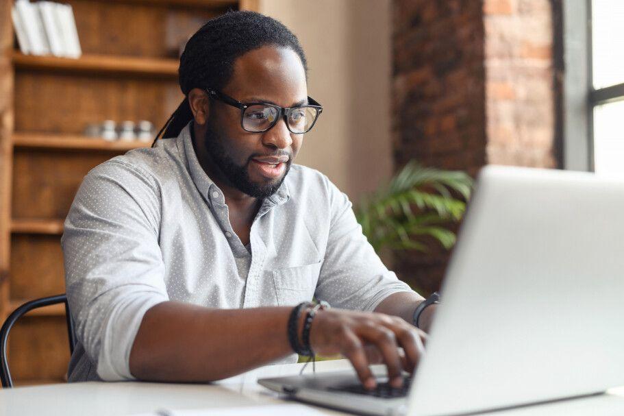 A professional man is seated at a desk, focused on his laptop. He is wearing glasses and a light-colored button-down shirt, typing attentively. The background includes a wooden bookshelf and a brick wall, suggesting a cozy and productive workspace.