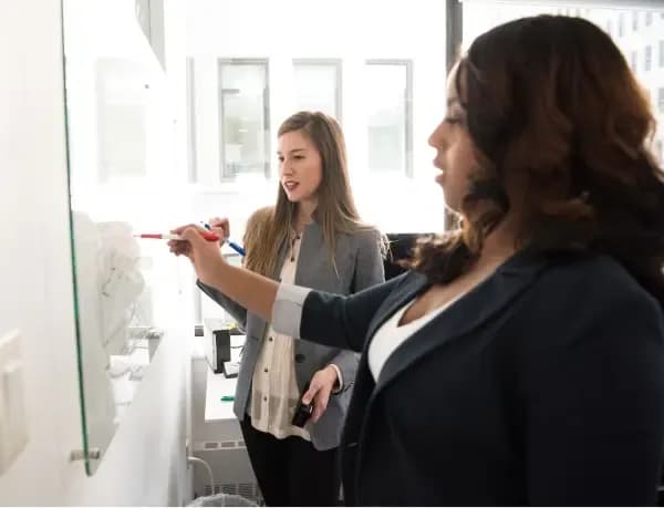 Two professional women in business attire collaborating and writing on a whiteboard in a bright office setting.