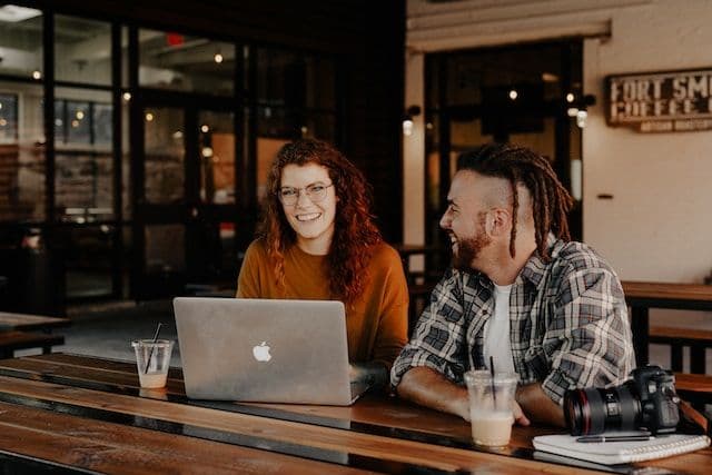 A smiling woman and a man with dreadlocks are sitting at an outdoor table with drinks, working together on a laptop. They appear to be in a casual, relaxed environment, suggesting a friendly and collaborative work setting.